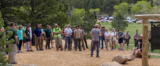 A large group of people gathered outdoors in a semi-circle listening to a speaker in front of a forest.