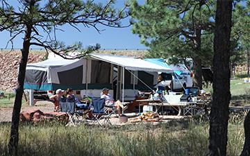A pop-up camper tent pitched at a sunny campsite surrounded by pine trees, with people sitting in lawn chairs outside.